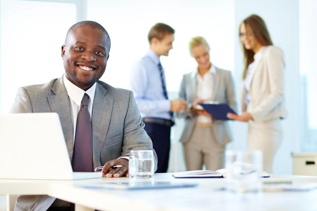 Professionnel souriant à son bureau, collègues en discussion à l'arrière-plan